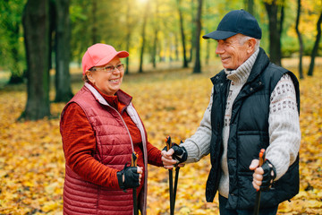 Pretty senior couple standing with nordic walking poles in colorful autumn park. Mature woman and old man resting outdoors.