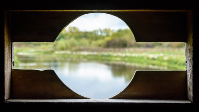 View from a birdwatching wooden hide over a pond through an opening with round shape cut-out to fit telephoto lenses. Sharp foreground (window), blurry background (landscape).