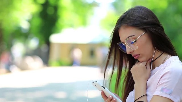 Attractive woman in glasses with smartphone and earphones listens to music in park. Beautiful woman with smartphone listens to music