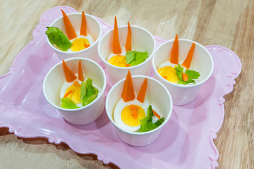 Food samples in small paper tasting cup on vintage pink tray. Cute decorative rabbit from boiled egg and vegetable