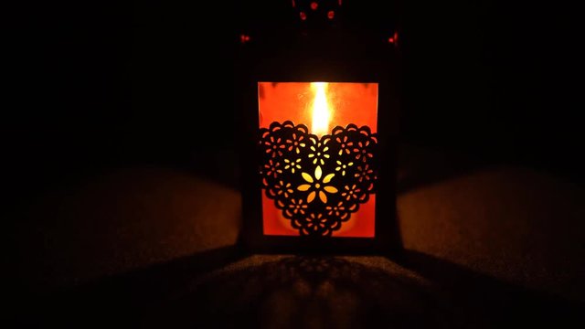 A yellow candle burns in a metallic red lantern with a delicate heart on a black background, close-up.