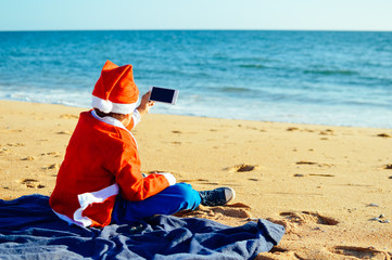 Fototapeta na wymiar Happy cute Santa child holding mobile smartphone showing blank display for your text message on light blue sky background. Happiness shopping time and electronic education lifestyle. Close up photo