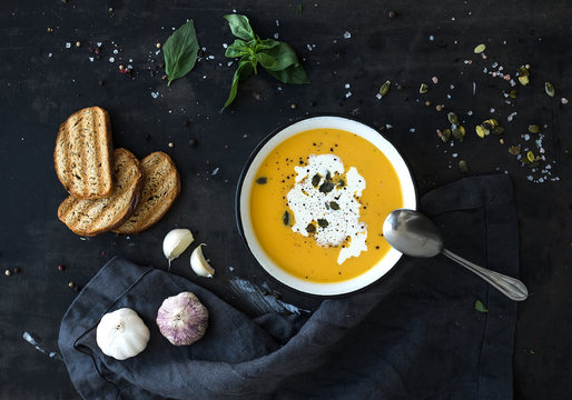 Pumpkin soup with cream, seeds, bread and fresh basil in rustic metal plate on grunge black background. Top view