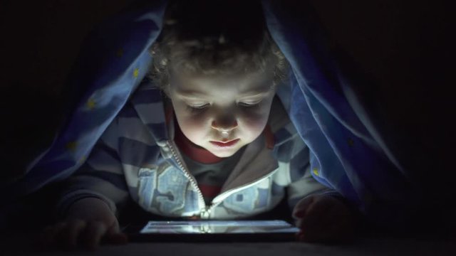 Little child, with blue eyes, watching a digital tablet under the bed sheets