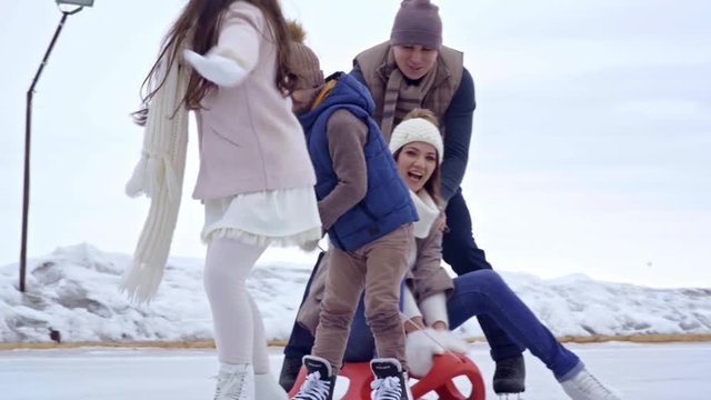 Happy children and cheerful father skating on ice and pulling sled with laughing young woman sitting on it while having fun in outdoor rink in winter