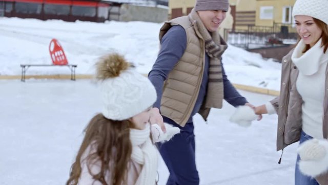 Tilt up of ecstatic man and woman holding hands with little children and turning in circle while ice skating outdoors, then spinning and laughing