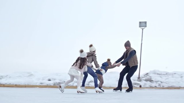Young man in warm puffy jacket and hat pulling happy woman and children skating in train formation on outdoor ice rink