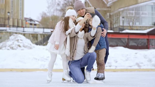 PAN of happy mother holding mobile phone and taking selfie with cheerful husband and cute children in skates posing on outdoor ice rink in winter