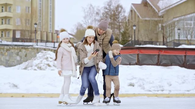 PAN of happy mother taking selfie with husband and cute little children after ice skating on outdoor rink in winter