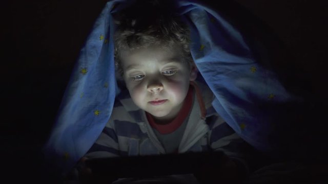 Little child, with blue eyes, watching a digital tablet under the bed sheets