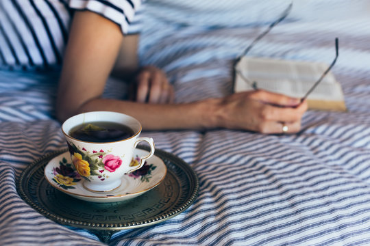 Woman reading book on bed with black tea in china cup next to her