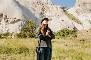 Naklejka premium Beautiful young woman in hat and gray sweater meditates with folded hands and closing eyes against backdrop of beautiful mountains and hills on sunny day
