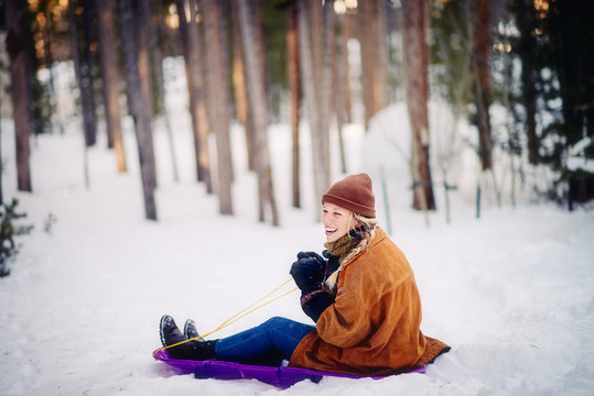 Teenage girl with a big smile sitting on a purple sled in snow amongst pine trees