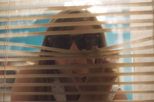 Young woman wearing sunglasses looks through blinds out a window