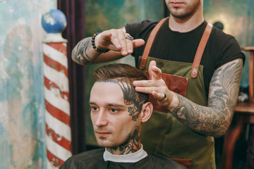 Close up shot of man getting trendy haircut at barber shop. Male hairstylist in tattoos serving...