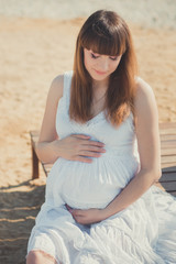 Genuine cute pregnant lady woman in white airy dress sitting sand beach wooden palette bridge holding tummy abdomen. Attractive beautiful young girl enjoying warm summer with unborn child in belly.