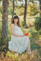 Adorable cute pregnant lady woman in white airy dress posing close to tree in forest holding tummy abdomen dreaming. Attractive beautiful young girl enjoying warm summer with unborn child in belly.