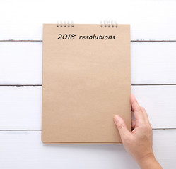Hand holding recycle paper notebook with 2018 resolutions over white wood background, sucess in business concept
