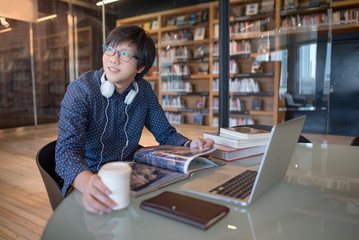 Young Asian man student reading book and using laptop computer in public library, education research and self learning in university life concepts