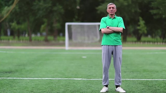 Football trainer stands on football field, physical education teacher. Senior man in green t-shirt stands on soccer field and looks at camera