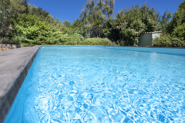 Surface of an empty swimming pool in a garden with clear cyan blue water, a small garden house and  bushes in the background