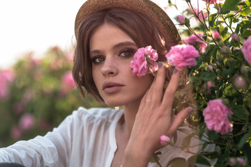 Fototapeta premium Beautiful young woman with curly hair posing near roses in a garden. The concept of perfume advertising.