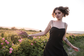 Beautiful young woman with curly hair posing near roses in a garden. The concept of perfume advertising.