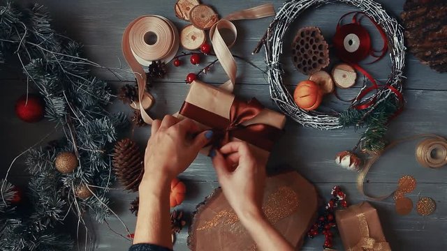 A top-down plan. Fully visible the table with the decorations. Female hands put and finalize Christmas gift wrapped in craftool paper on a wooden table. Bandaging tape and tied in a bow.