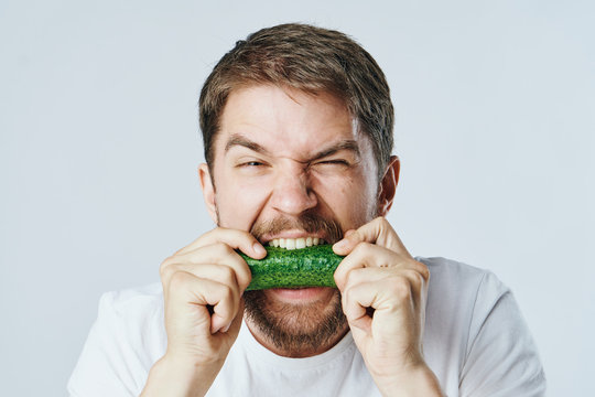 A Man With A Beard And A White T-shirt Biting Into A Cucumber