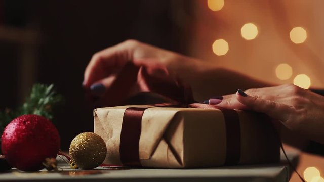 Close-up on the background of Christmas lights female hands open a gift from Kraft paper. The background is blurred in bokeh.