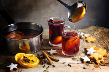 pouring mulled wine in glass mugs and a pot, Christmas cookies and spices on a rustic wooden table,...
