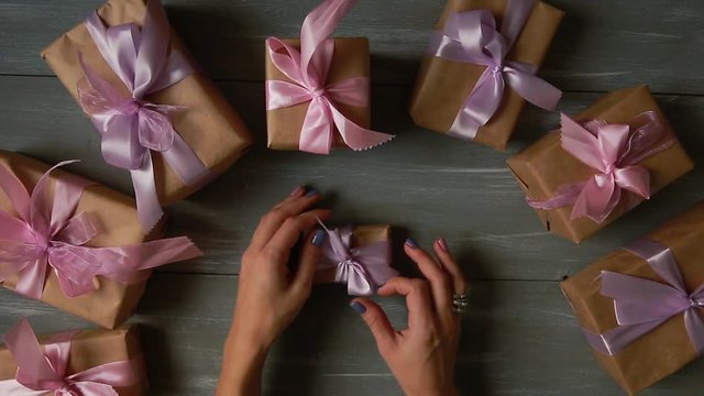 A top-down plan. Fully visible the table with the decorations. Female hands put and finalize Christmas gift wrapped in craftool paper on a wooden table. Bandaging tape and tied in a bow.