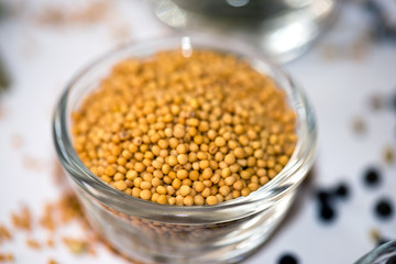 A close-up view of a glass bowl filled with yellow mustard seeds, surrounded by scattered mustard seeds and black peppercorns on a white surface, highlighting the texture and natural appearance