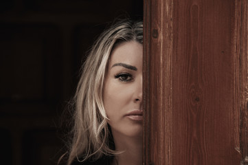  Beautiful girl peeks through the door, peeking Out of Doorway. GIrl with low lighting, a massive door covers half face closeup. Suspicious and attentive look.