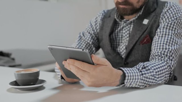 Front view on unrecognizable bearded business man in casual wear working on his touchscreen smart tablet , sitting in industrial well lita cafe shop at morning, next to cup with flat white coffee