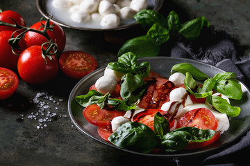 Italian caprese salad with sliced tomatoes, mozzarella cheese, basil, olive oil. Served in vintage metal plate with ingredients above over dark metal background. Close up. Rustic style - obrazy, fototapety, plakaty