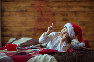 Obraz premium Girl child in jersey in santa hat on the bed, lifestyle, real interior, rustic style, red and brown wooden background