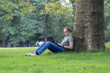 Naklejka na ściany i meble Young man in glasses work on laptop sitting on the grass near the tree in park. Maybe student or hacker businessman