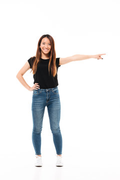 Full length photo of happy charming asian woman in casual wear standing with outstretched hand, pointing with finger, looking at camera