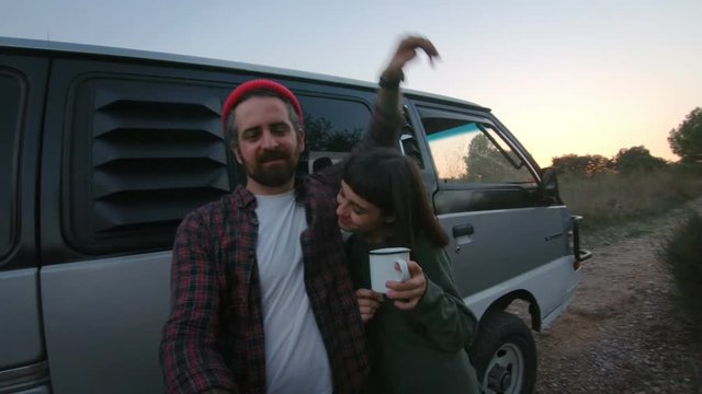 Cute hipster couple of young millennials make video or photo selfie during trip in camping caravan or van, stand in front of car, drink tea and laugh, emjoy youth and life in nature