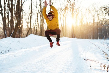 Handsome young runner in sportswear with headphones jumping on snow covered winter road in the morning near the forest.