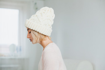 Portrait of young woman in white knitted hat. Sad girl indoors in warm clothes.