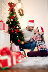 Young pretty happy girl hugging her handsome boyfriend while he looking her with a smile on the floor at home for Christmas holidays.