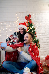 Young adorable smiling husband and wife sitting on the floor with a blanket and taking a selfie for Christmas.