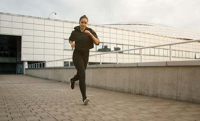 Fototapeta na wymiar Young attractive lady jogging in the city near buildings. Beautiful happy girl running in the morning outside. Woman trains during the sunrise in the city center before breakfast. 