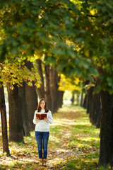 A beautiful happy smiling brown-haired woman in white sweater standing with a red book in fall city park on a warm day. Autumn golden leaves. Reading concept.