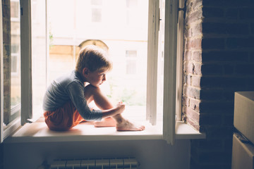 Sad preschooler boy sitting on windowshill. Childhood, relocation loneliness concept.