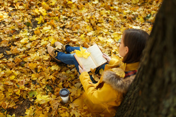 A beautiful happy brown-haired woman in yellow coat and jeans sitting under the maple tree with cup...