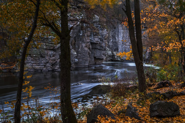 The stream of water in the river flowing between the rocks in the canyon. Long exposure. Trees with golden leaves on the river bank.

