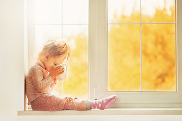 Little cute Kid Girl sitting by window indoor holding cup of hot drink cocoa enjoying autumn forest background. Season Beauty fashion Children concepts. Fall season